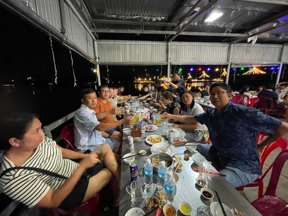 A long table of family and friends at a riverside restaurant at night, glasses raised in a toast, with city lights reflected on the water behind them